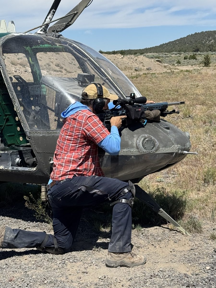 James McNames aiming a precision rifle braced across the nose of a helicopter on a high-desert range